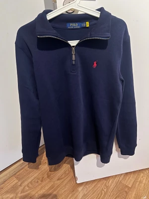 Polo Ralph Lauren tröja - Mörkblå stickad tröja från Polo Ralph Lauren med halv dragkedja och klassisk röd logga broderad på bröstet. Tröjan har hög krage, ribbade muddar och är tillverkad i mjukt material. Perfekt för lager på lager och en stilren look.