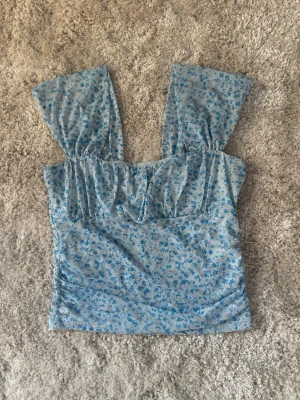 Blommig blå linnetopp med breda axelband - Söt ljusblå linnetopp med småblommigt mönster i blått. Toppen har breda axelband och en lätt rynkad detalj vid bysten. Perfekt för varma dagar och ger en romantisk vibe till din outfit.