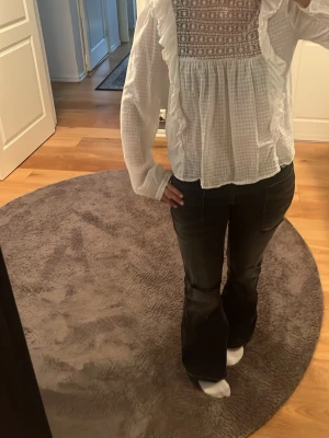 Vit blus med spets och volanger - Söt vit blus med långärmad design, spetsdetaljer upptill och volanger längs bröstet. Blusen har ett rutigt mönster i tyget och en lös, luftig passform. Perfekt för dig som gillar romantisk och trendig stil.