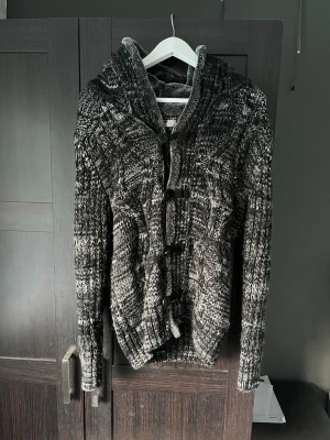 Gråmelerad stickad cardigan med luva - Säljer en riktigt schysst cardigan i et otrolig fint färgmönster! Fodrad i luvan och längs med knapparna. Skriv vid funderingar!