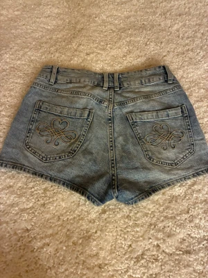 Blå denimshorts från Lindex - Snygga blå denimshorts från Lindex med medelhög midja och broderade detaljer på bakfickorna. Klassisk femficksmodell med bälteshällor och dubbla knappar framtill. Perfekta för varma dagar och enkel att matcha med olika toppar.
