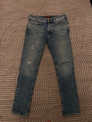 Blå jeans från Jack & Jones med slitningar - Säljer ett par blå skinny jeans från Jack & Jones med slitningar på låren och knäna. Jeansen har klassisk femficksmodell, normal midja och är tillverkade i stretchigt denimtyg för skön passform. Perfekta för en avslappnad och trendig look.
