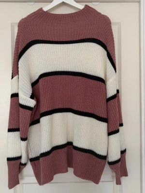 Randig oversized stickad tröja 1XL - Säljer en oversized stickad tröja i dammig rosa, vit och svart. Tröjan har breda horisontella ränder, ribbstickad halsringning och muddar. Perfekt för lager på lager och en avslappnad look.