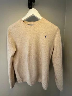 Beige stickad tröja från Polo Ralph Lauren - Klassisk beige stickad tröja från Polo Ralph Lauren med rund halsringning och diskret blå logga broderad på bröstet. Tröjan har ribbade muddar vid ärmslut och nederkant och är tillverkad i mjukt material som passar perfekt till höst och vinter. Nypris 1750 kr