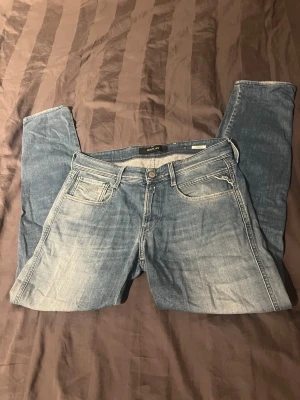 Blå jeans från Replay - Säljer ett par snygga blå jeans från Replay med klassisk slim modell och slitna detaljer. Jeansen har en tvättad look. Perfekta för dig som gillar en avslappnad men trendig stil.