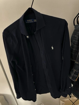Mörkblå skjorta från Polo Ralph Lauren - Klassisk mörkblå skjorta från Polo Ralph Lauren med vit broderad logga på bröstet. Skjortan har lång ärm, knappar framtill och är tillverkad i mjuk bomull. Storlek XS