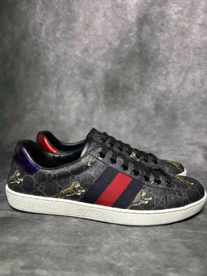 Gucci Ace monogram  - Svarta Gucci Ace sneakers i läder med GG-monogram, röda och blå ränder på sidan samt tigerdetaljer. Skon har vit sula, svarta skosnören och kontrasterande hälflikar i rött och lila. Perfekt för dig som vill sticka ut med exklusiv streetstyle.