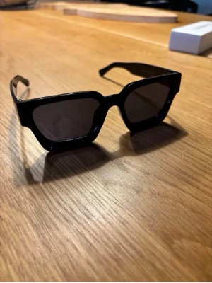 Svarta kantiga solglasögon - Snygga solglasögon med bred, svart båge och mörka glas. De har en trendig, kantig form som ger en cool och modern vibe. Perfekta för dig som vill sticka ut och addera edge till din stil.