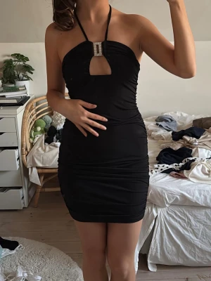 Svart halterneck kortklänning - Säljer en svart kort klänning med halterneck och dekorativt smycke framtill. Klänningen har en tight passform och en liten cut-out vid bysten. Perfekt för studentresor, sommaren.