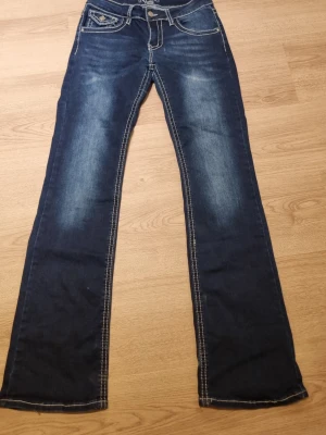 Mörkblå bootcut jeans med kontrastsömmar - Snygga mörkblå bootcut jeans med tydliga kontrastsömmar. Jeansen har klassiska dekorativa bakfickor med knappdetaljer. Tillverkade i denim med lätt stretch för skön passform.