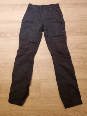 Svarta byxor med dragkedjeficka - Svarta byxor i smal passform med flera fickor, inklusive en dragkedjeficka på låret. Förstärkta knän och justerbara benslut med tryckknappar ger en funktionell look. Tillverkade i slitstarkt tyg, perfekta för dig som gillar praktiska detaljer. Friluftsbyxor. 