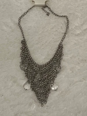 Silvrigt halsband med kedjor och kristaller - Unikt halsband i silvrig metall med flera lager av kedjor som bildar en trekantig form. Dekorerat med stora, klara kristalliknande hängen som ger en cool statement-look. Perfekt för dig som vill sticka ut med accessoarerna.