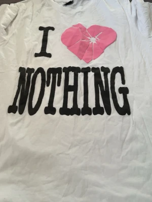 Vit t-shirt med tryck och trasigt hjärta - Vit t-shirt med svart texten 'I NOTHING' och ett rosa trasigt hjärta på bröstet. T-shirten har korta ärmar och är gjord i mjuk bomull. Perfekt för dig som gillar statement-plagg med attityd.