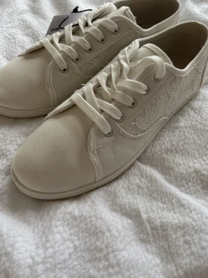 Vita spetsdetaljerade sneakers - Snygga vita sneakers. Skorna har klassisk låg modell, vita snören och ovandel i textil. Perfekta för dig som vill ha en stilren men unik look till vardagsoutfiten.