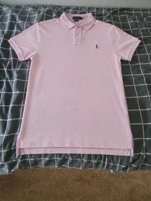 Ljusrosa pikétröja från Ralph Lauren - Snygg ljusrosa pikétröja från Ralph Lauren med klassisk krage och två knappar framtill. Tröjan har korta ärmar och den ikoniska broderade loggan på bröstet. Perfekt för en stilren och avslappnad look. Specialgjord i USA. Pris kan sänka vid snabbt köp