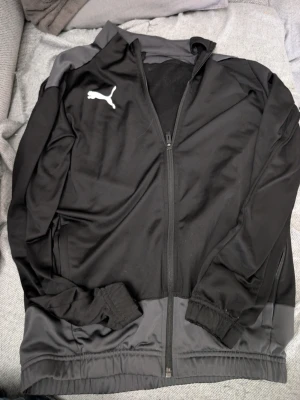 Svart träningsjacka från Puma stl 152 - Snygg svart träningsjacka från Puma med dragkedja framtill och vita Puma-loggor på bröstet och etiketten. Jackan har ribbade muddar, grå detaljer på axlarna och är tillverkad i ett mjukt, sportigt material som passar perfekt till träning eller chill.