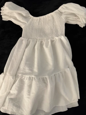 Vit kort klänning med puffärm - Söt vit kortklänning med puffiga kortärmar off shoulder och smockad överdel. Klänningen har volangdetaljer och är tillverkad i ett lätt, luftigt material som passar perfekt för varmare dagar. Stilren och trendig design.