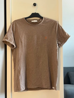 Brun t-shirt från Les Deux - Snygg brun t-shirt från Les Deux med liten broderad logga på bröstet. Klassisk rund halsringning och korta ärmar. Tillverkad i mjuk bomull för en skön känsla. Perfekt till jeans eller shorts för en avslappnad stil.