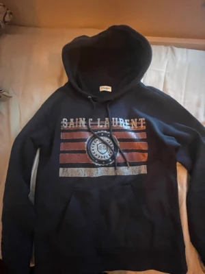 Mörkblå Saint Laurent hoodie perfekt skick - Mörkblå hoodie från Saint Laurent med stor huva och dragsko. Framsidan har ett coolt tryck med texten 'SAINT LAURENT' och grafiska ränder i rött och vitt samt en rund logga i mitten. Perfekt för dig som gillar streetstyle och vill sticka ut. Storlek xs längd 58 bredd 45 cm. Kan gå ner i pris lite😁👌