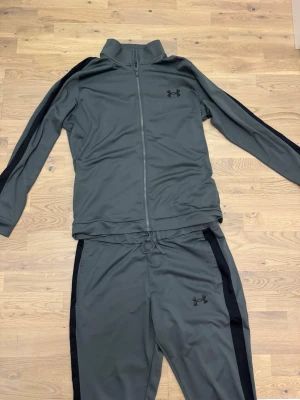 Grått träningsset från Under Armour - Snyggt grått träningsset från Under Armour med svarta detaljer längs ärmar och ben. Setet består av en zip-jacka med hög krage och matchande byxor med dragsko i midjan. Perfekt för gymmet eller sportaktiviteter.