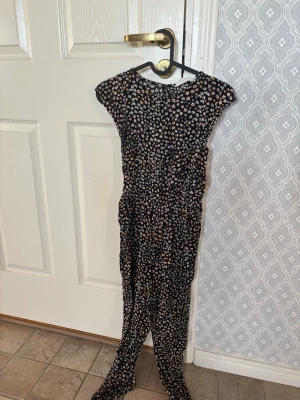 Blommig svart byxdress med kort ärm - Svart byxdress med små blommor i vitt, rosa och gult. Kort ärm och knäppning i nacken. stl 146.