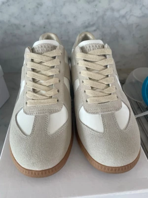 Beige och vita sneakers med mockadetaljer - Stilrena sneakers i beige mocka och vitt läder med klassisk gummisula. Snygga detaljer och neutrala färger gör dem lätta att matcha. Perfekta för dig som gillar minimalistisk och trendig stil.