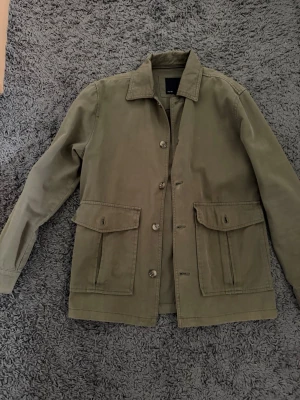 Olivgrön overshirt med fickor - Stilren olivgrön overshirt med klassisk krage och knäppning framtill. Två stora fickor med lock på framsidan ger en cool utility-vibe. Tillverkad i ett kraftigare bomullstyg som passar perfekt till lager-på-lager-stil.