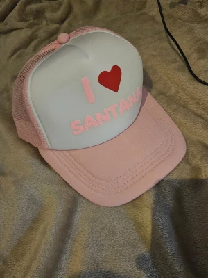 Papi santana Rosa och vit truckerkeps I ❤️ SANTANA - Säljer en trendig truckerkeps i rosa och vitt med texten 'I ❤️ SANTANA' framtill. Kepsen har mesh på sidorna och bak, justerbar rem och böjd skärm. Perfekt för dig som vill sticka ut med en färgglad accessoar.