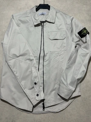 Stone Island - Snygg vit overshirt från Stone Island med klassisk krage, dragkedja framtill och bröstficka med lock. Ikonisk svart patch med logga på vänster ärm. Tillverkad i ett slätt, lätt material som ger en clean look.