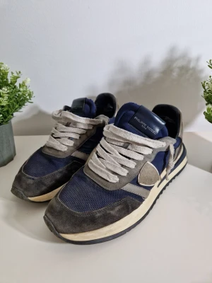 Philippe Model blå och grå sneakers - Snygga sneakers från Philippe Model i marinblått och grått med vita detaljer. Ovandelen är i en mix av mocka och mesh, med breda vita skosnören och en klassisk låg siluett. Skon har en vit och blå randig sula och logga på sidan.