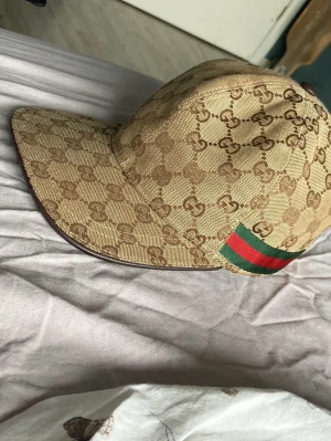 Gucci keps äkta  - Snygg beige keps från Gucci med klassiskt GG-monogram över hela kepsen och en grön-röd rand på sidan. Kepsen är i storlek L (59 cm) och har en böjd skärm. Materialet är canvas med bomullsfoder på insidan. Perfekt för dig som vill sticka ut med en lyxig accessoar.