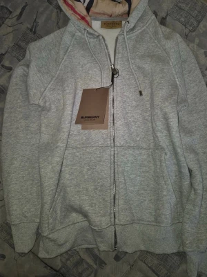 Grå zip-hoodie från Burberry - Snygg grå hoodie från Burberry med dragkedja och klassisk huva. Insidan av huvan har det ikoniska Burberry-mönstret. Tröjan har känguruficka framtill och ribbade muddar vid ärmslut och nederkant. Perfekt för en stilren och avslappnad look.