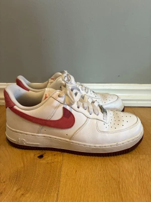 Nike Air Force 1 vita med rosa swoosh - Klassiska Nike Air Force 1 sneakers i vitt läder med rosa swoosh och yttersula. Lågt skaft, perforerad tåbox och vita skosnören. Ikonisk modell med bekväm passform och stilren design som passar till många outfits. Använda endast ett fåtal gånger.
