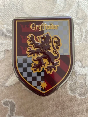 Gryffindor plåtask Harry Potter - Snygg plåtask i form av Gryffindors vapensköld från Harry Potter. Asken är röd, gul och silver med ett lejonmotiv och texten 'Gryffindor' på locket. Perfekt för småsaker eller samlare av Harry Potter-prylar.