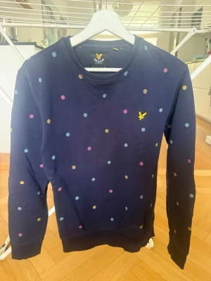 Mörkblå prickig tröja Lyle & Scott - Mörkblå sweatshirt från Lyle & Scott med färgglada prickar. Tröjan har rund halsringning och klassisk gul logga broderad på bröstet. Perfekt för dig som gillar en lekfull och stilren look.