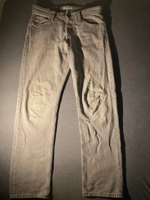 Grå jeans från Jack & Jones - Snygga grå jeans från Jack & Jones med klassisk femficksdesign och raka ben. Jeansen har en läderpatch med logga bak i midjan och är tillverkade i bomull. Perfekta för en avslappnad och stilren look.