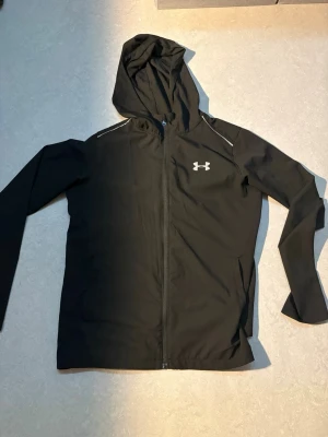  Under Armour - Svart träningsjacka med huva från Under Armour. Jackan har hel dragkedja, två fickor fram och vit logga på bröstet samt stort Under Armour-tryck på ryggen. Lätt och smidig modell, perfekt för löpning eller gym. Materialet är troligtvis polyester.