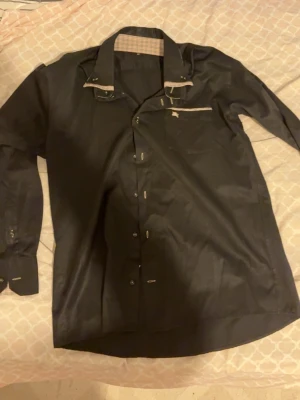 Svart skjorta med rutiga detaljer - Snygg svart skjorta från Burberry med klassiska rutiga detaljer på insidan av kragen och fickan. Skjortan har lång ärm, knappstängning och en liten broderad logga på bröstfickan. Perfekt för dig som gillar stilrena och exklusiva plagg.
