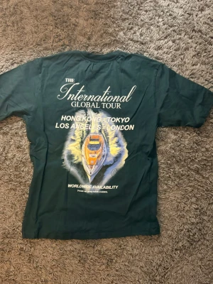Mörkgrön t-shirt International Tour - Mörkgrön t-shirt med tryck på både fram- och baksida. På ryggen finns ett stort motiv med båt och text om 'The International Global Tour' samt städer som Hong Kong, Tokyo, Los Angeles och London. Rund hals och korta ärmar.