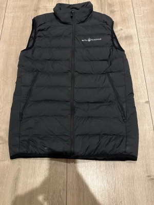 Svart dunväst från Sail Racing - Snygg svart dunväst från Sail Racing med diskret logga på bröstet. Västen har hög krage, två dragkedjeförsedda fickor och quiltad design. Perfekt för lager-på-lager och funkar till de flesta outfits. Materialet är dun och polyester.