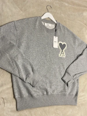 Grå sweatshirt från Ami Paris - Grå sweatshirt från Ami Paris med stort hjärtformat A-märke på bröstet. Tröjan har rund hals, ribbade muddar och är gjord i mjukt bomullsmaterial. Perfekt för en avslappnad och trendig look. Skriv gärna till mig för mer information samt detaljer. Mvh Seller