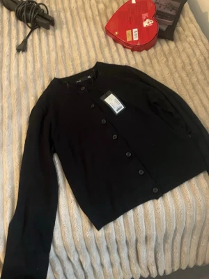 Svart kofta  - Stilren svart kofta från gtab.1999 med rund halsringning och knäppning framtill. Koftan har långa ärmar och ribbstickade detaljer vid halsen. Perfekt att slänga över en t-shirt eller klänning för en enkel och clean look.