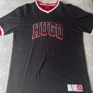 Svart t-shirt från Hugo med tryck - Svart t-shirt från Hugo med röd och vit V-ringad krage och ärmslut. Stor HUGO-logga i rött och vitt framtill samt patch med siffran 93 nertill. Perfekt för en sportig och trendig look.
