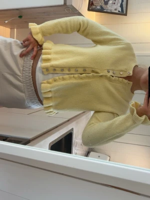 Gul cropped kofta med volangkant - Söt ljusgul cropped kofta med volangkant och små knappar framtill. Koftan har långa ärmar och är stickad i ett mjukt material. Perfekt för att liva upp din outfit med en poppig färg.