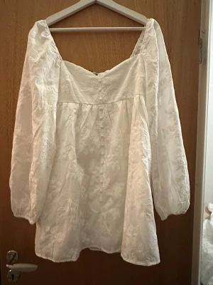 Vit broderad klänning från Gina Tricot - Superfin vit klänning från Gina Tricot med broderade blommor och knappar framtill. Klänningen har långärmade puffärmar och en söt v-ringning. Perfekt för sommarens alla tillfällen!