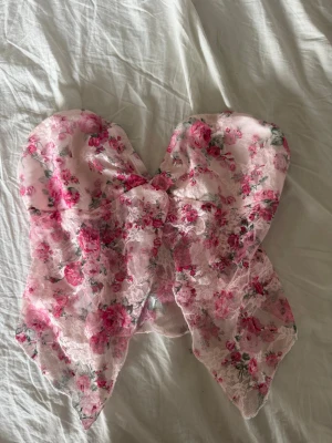 Blommig axelbandslös tubtopp i spets - Supersöt axelbandslös tubtopp med blommigt mönster i rosa och vitt. Toppen har en stor rosett framtill och är gjord i genomskinlig spets som ger en romantisk vibe. Perfekt för dig som vill sticka ut med en unik och feminin stil.