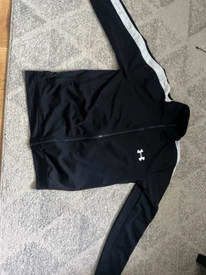 Svart träningsjacka från Under Armour - Svart träningsjacka med vit logga från Under Armour. Jackan har hel dragkedja, ståkrage och vita ränder längs ärmarna. Tillverkad i ett slätt och sportigt material som passar perfekt till träning eller uppvärmning.