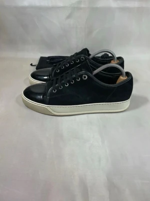 Svarta sneakers från Lanvin - Stilrena svarta sneakers från Lanvin med ovandel i mocka och detaljer i blankt läder på tån och hälen. Skorna har vita sulor och svarta skosnören, samt metallfärgade öljetter. Perfekta för en clean och modern look. Pris kan diskuteras😊