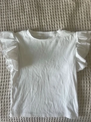 Vit topp med volangärmar - Superfin vit ribbad topp med volangärmar från gina tricot. Toppen har rund halsringning och volanger på ärmarna. Knappast använd!