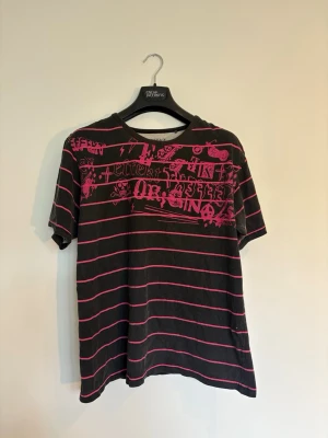 Svart och rosa randig t-shirt - Cool svart t-shirt med rosa ränder och tryck framtill. T-shirten har korta ärmar och en avslappnad passform. Perfekt för dig som gillar en edgy och unik stil.